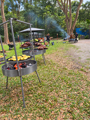 Outdoor campsite scene with multiple round metal charcoal grills on a grassy lawn, some with food cooking and others empty. Trees, smoke, and a few people are visible in the background.