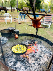 Outdoor cooking scene with roasted meats and vegetables over a fire pit, surrounded by chairs and horses.