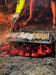 A folding BBQ hot plate being used over an open fire, cooking sausages and skewers.