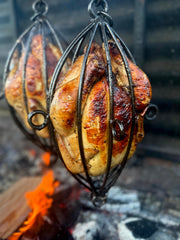 Two roasted chickens hanging over an open flame on a metal rack.