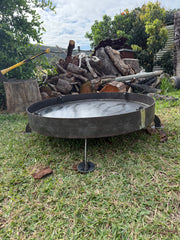 A round metal fire pit with a black finish and three legs, filled with water, set on a grassy lawn with a pile of firewood and trees in the background.