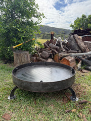 A round black metal fire pit with three legs sits on grass outdoors, with a pile of logs, an axe, and trees in the background under a cloudy sky.