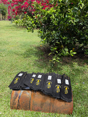 Several black protective gear sleeves or wraps with yellow printed logos are laid on top of a rusted metal barrel in a grassy outdoor area with flowering bushes in the background.
