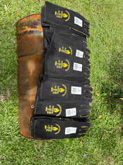 A stack of black leather work gloves with yellow logo prints and white labels, laid out on a rusty metal barrel on a green grass background.
