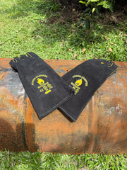Two black leather welding arm sleeves with yellow embroidery, displayed on a rusty metal barrel outdoors on a grassy lawn.