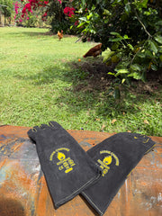 Two black fire-resistant protective sleeves with yellow ACK logos, displayed on a rusty metal surface with a grassy backyard and chickens in the background.