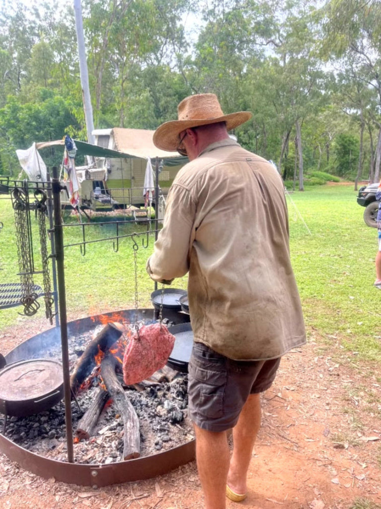 Man cooking over an open fire in a natural setting with trees and grass.