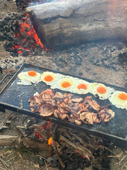 Rectangular outdoor hot plate cooking eggs with bright orange yolks and bacon over an open wood fire, with embers and ash visible around it.