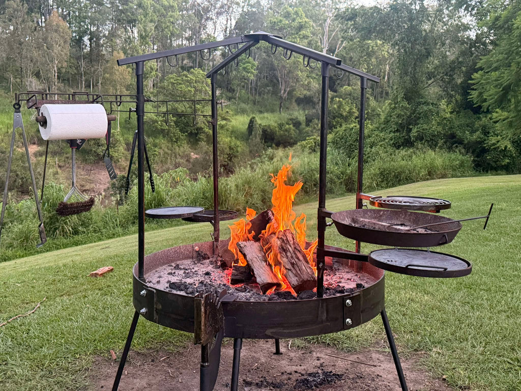 An outdoor fire pit with a metal roof and multiple cooking grates, set in a grassy area, with fire visible beneath the grates.
