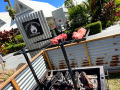 Outdoor grill with meat cooking over a fire pit, featuring 'Aussie Campfire Kitchens' sign.