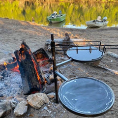 Outdoor camping cooking setup featuring a hot plate and grill with accessories over a campfire, surrounded by a body of water and boats in the background.